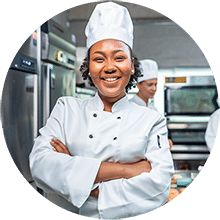 A happy chef with her arms crossed wearing all white in a commercial kitchen.